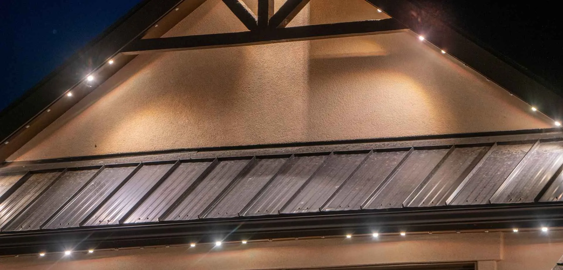 Close-up of a stucco gable with timber trusses and a metal roof edge, outlined by small white roofline lights at night